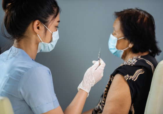 A healthcare provider administers a vaccine to an older woman. Both people are wearing face masks.